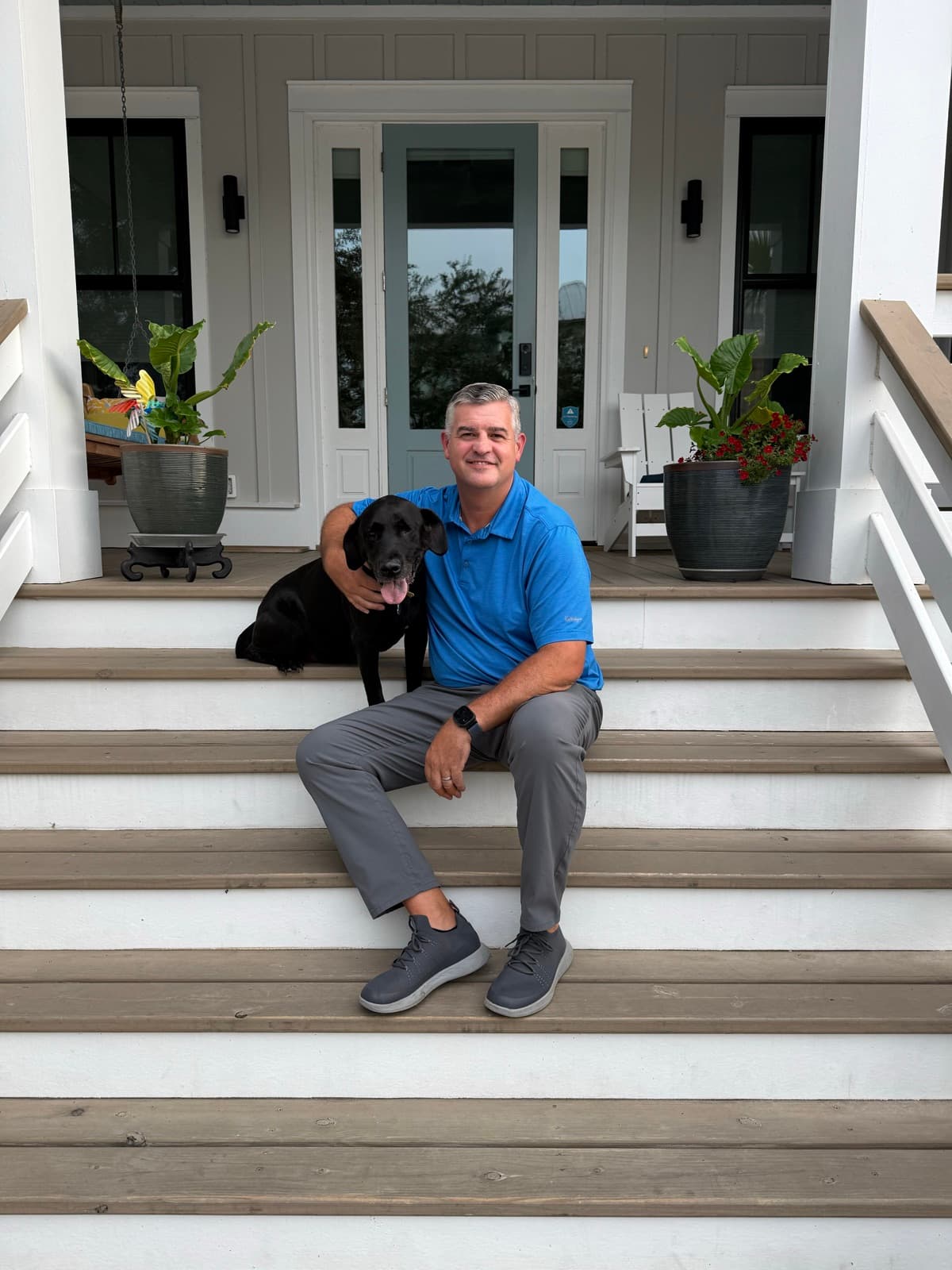 Chris Egan, founder of TideLine Overwatch, sitting on the front steps of a coastal home with his black Labrador